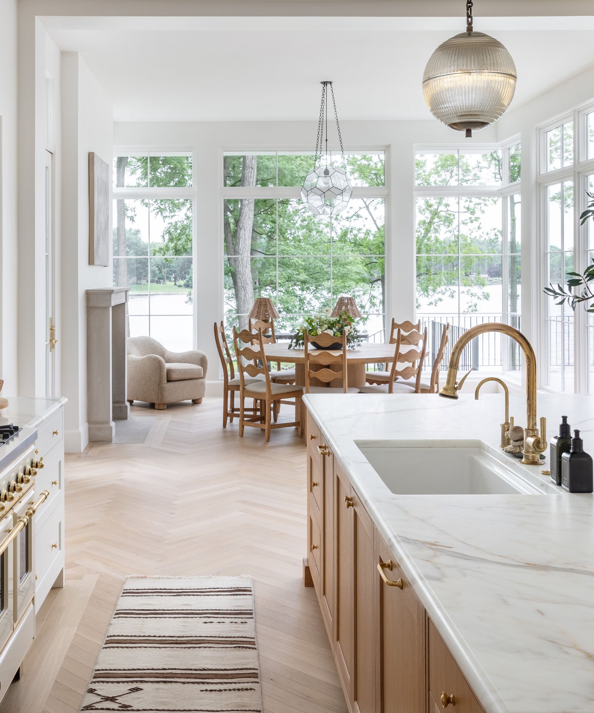 A wood and white kitchen with an eat-in area at the end, surrounded by floor to ceiling windows with views over a lake
