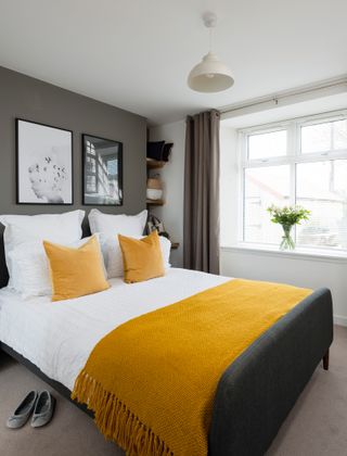A master bedroom in a grey colour scheme with double bed, mustard yellow throw, and shelving in the corner of the room