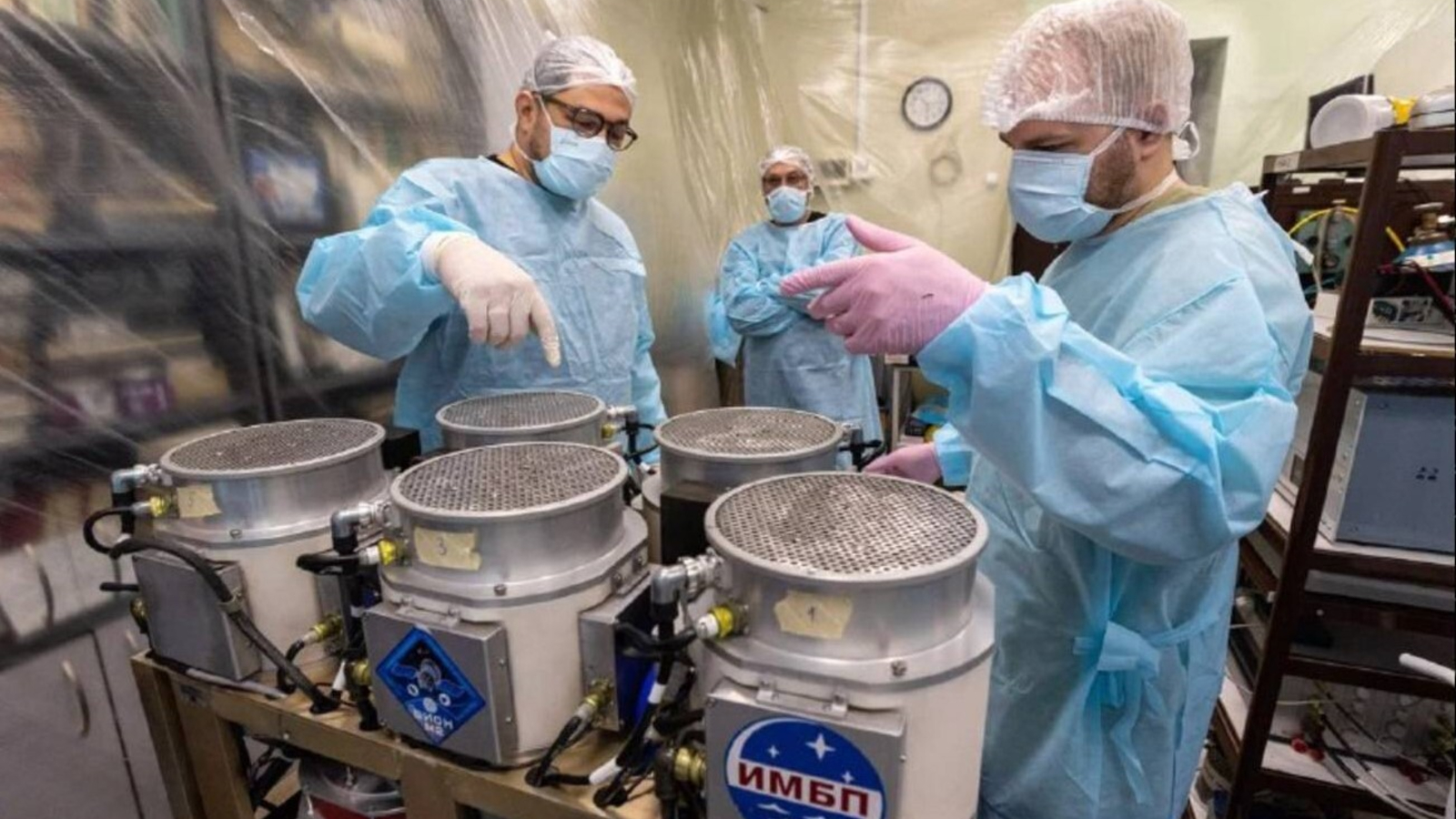 Three people wearing blue clean suits and hair nets with purple gloves stand next to a crate of metal drums in a cleanroom.