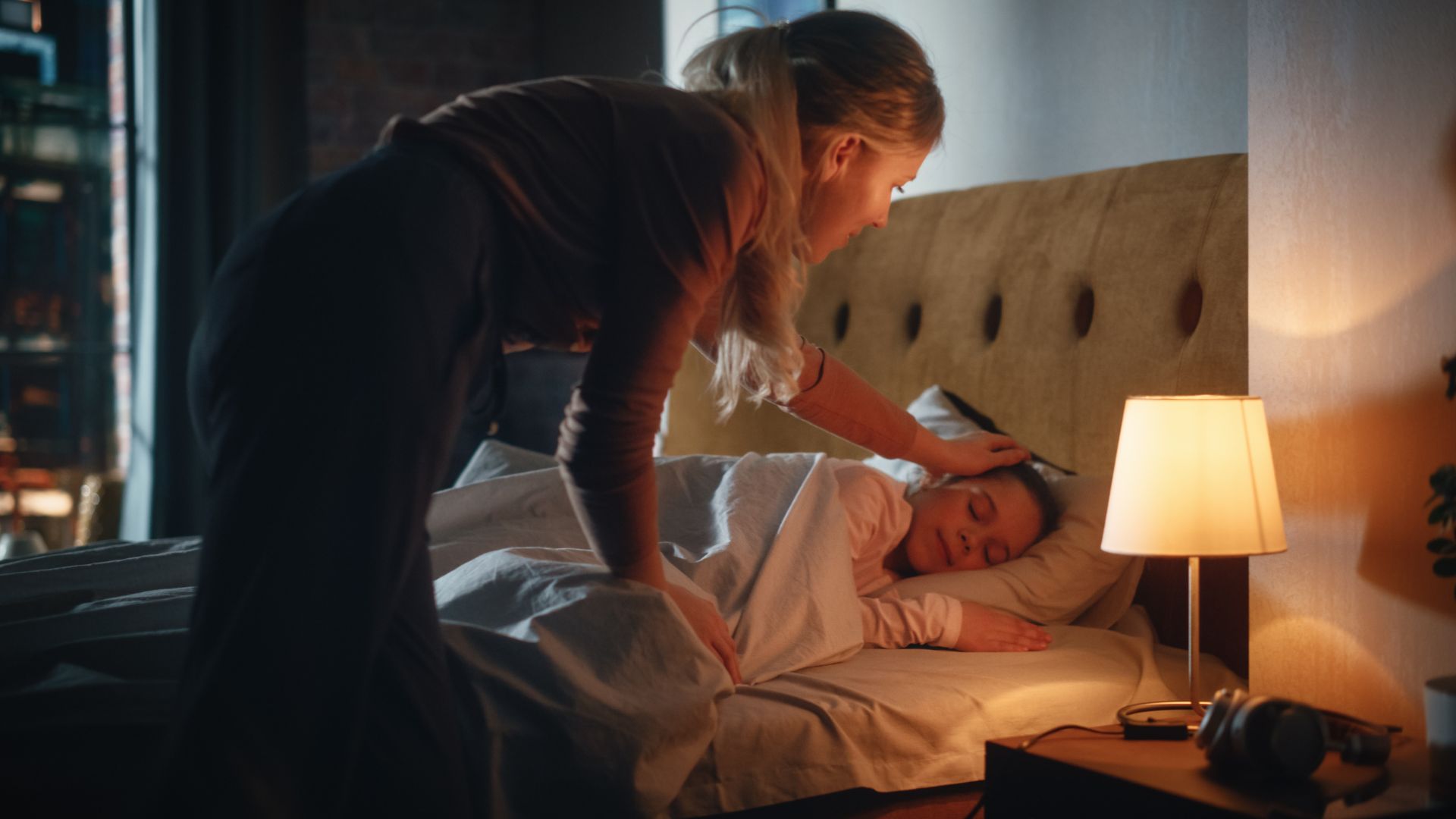 A mom strokes the head of her daughter as the daughter lies in bed in a dark bedroom with a nightlight on the bedstand nearby.