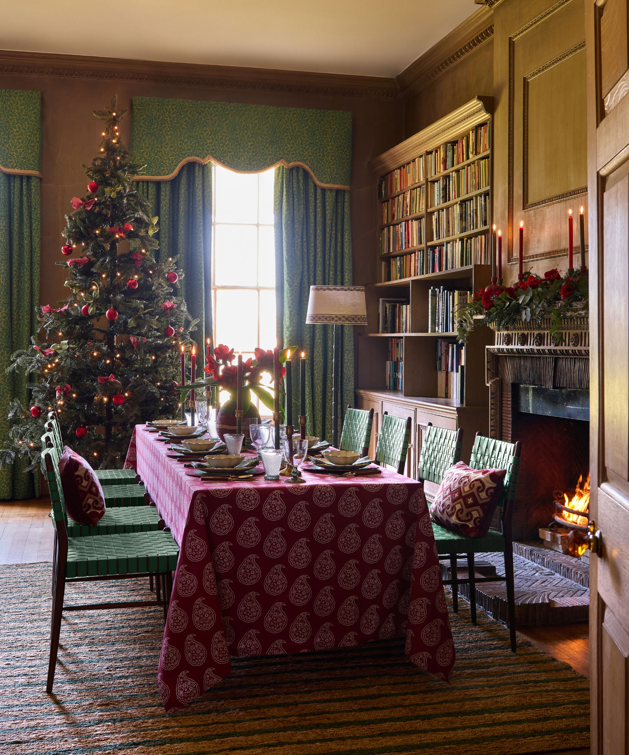 christmas dining room with wood panelled walls, a lit fireplace, striped rug with a dining table set with green chairs and a paisley tablecloth with a large christmas tree set behind