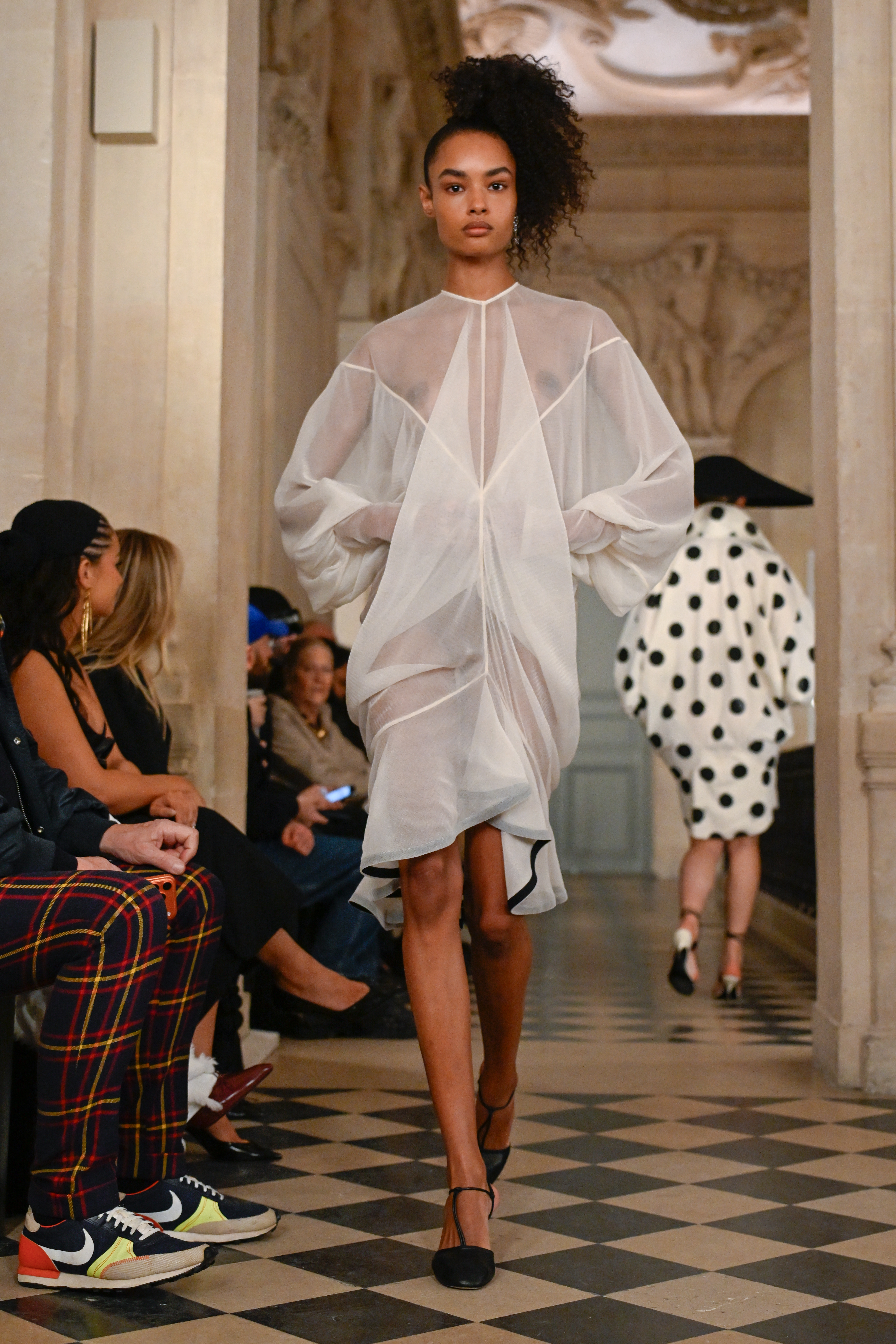 a model walks during a runway show wearing an oversized, white, mesh gown and black pumps. her curly hair is styled in a side ponytail