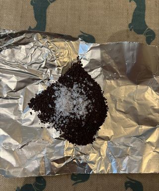 A pile of coffee grounds with salt sprinkled on top on a piece of tinfoil on a beige and turquoise dachsund printed table cloth