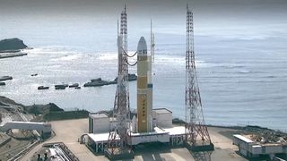 an orange rocket waits on a launch pad with a rocky ocean scene behind it