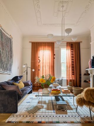 A light, modern, high ceiling living area with tall windows along the far wall, dressed with custom rust coloured curtains, and a white, industrial style chandelier. On the left wall hangs a monochrome textile piece, above a deep, plum coloured sofa. In the centre of the space is a geometric design rug, with a glass coffee table and modern, fur upholstered chair.