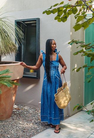 A woman's jean dress outfit with a maxi denim dress styled with a raffia tote bag and black flip-flops.