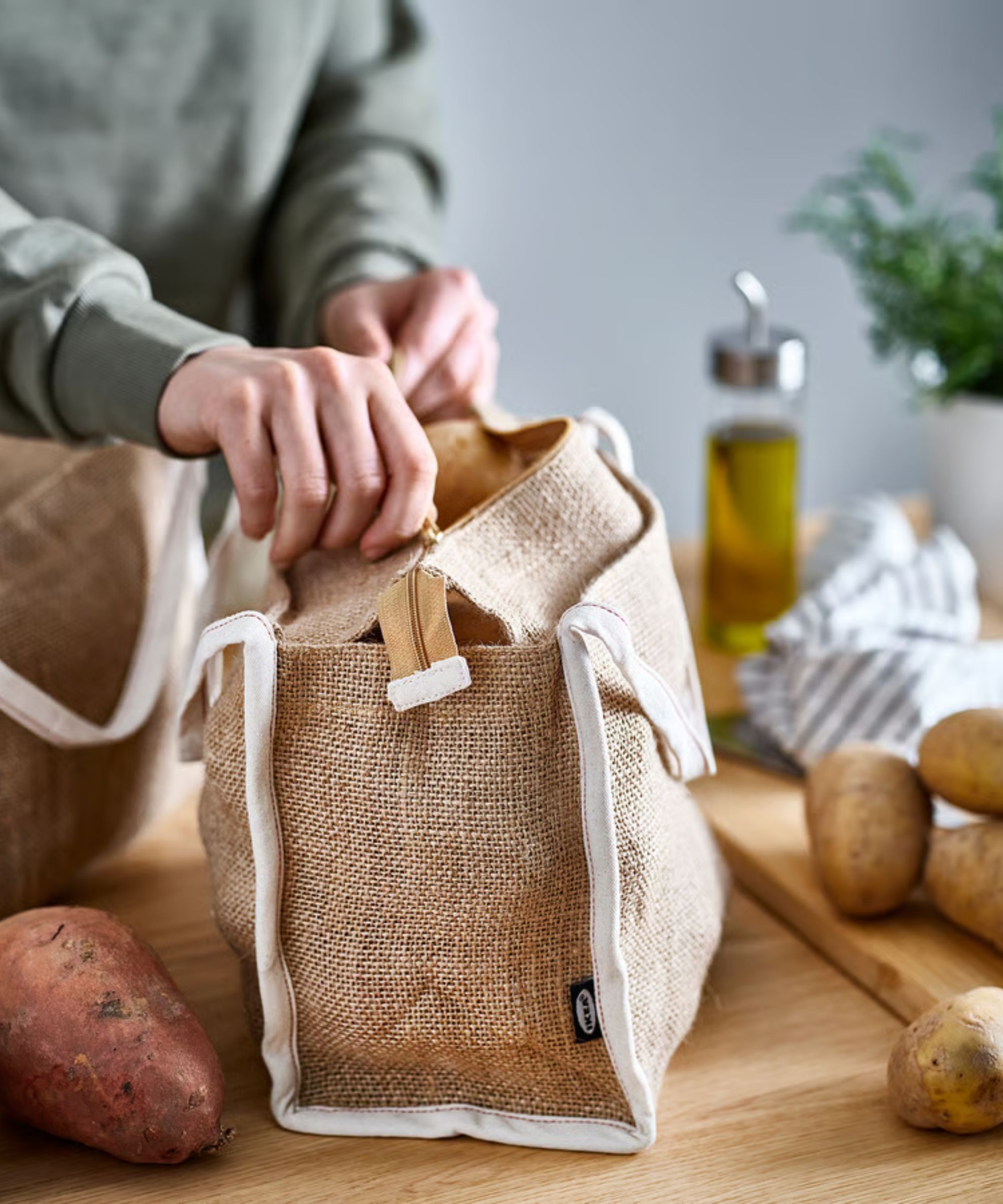 A Caucasian man's hands zipping up the top of a jute food storage bag on a wooden kitchen counter, surrounded by potatoes.
