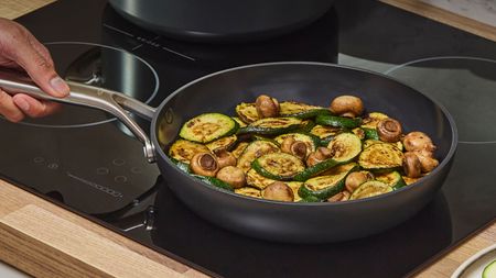 A black ceramic frying pan on a black stovetop with mushrooms and zucchini inside. 