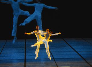 The male and female dancers dressed in all white are photographed mid-movement. There is a projection of them in the background.