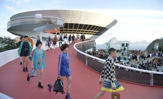 Models walking down a spiraling red walkway coming down from a saucer shaped building with a crowd of people below.