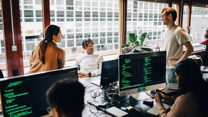 Mixed age group of cybersecurity professionals speaking in an open plan office space with colleagues working on desktop computers in foreground.