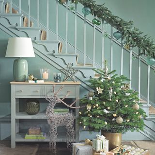 Wrapped gifts and christmas crackers next to gold pot with small tree and decorations in front of decorated green staircase