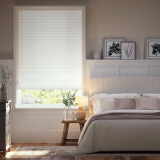 White blind covering a window in a cream bedroom with upholstered bed and neutral cushions