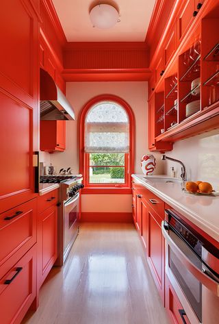 burnt orange galley kitchen with all units painted the same, wooden floor, white countertops, arched window with blind