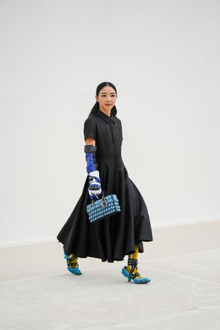 Yuwei Zhangzou wears a black short sleeves / long pleated dress from Dior, a black / blue / white shiny leather high biker gloves from Dior, a black with embroidered pale blue fringed / pearls East-West handbag from Dior, yellow mustard with white print pattern socks from Dior, black with embroidered pale blue fringed / pearls pointed / ankle ballerinas from Dior , outside Dior, during Paris Fashion Week - Womenswear Spring/Summer 2023, on September 27, 2022 in Paris, France