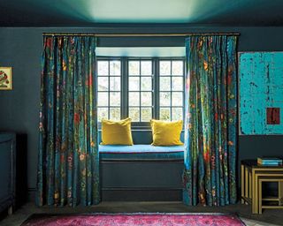 Window seat with blue and yellow cushions and colourful floral curtains. Nest of tables. Modern artwork in blue.