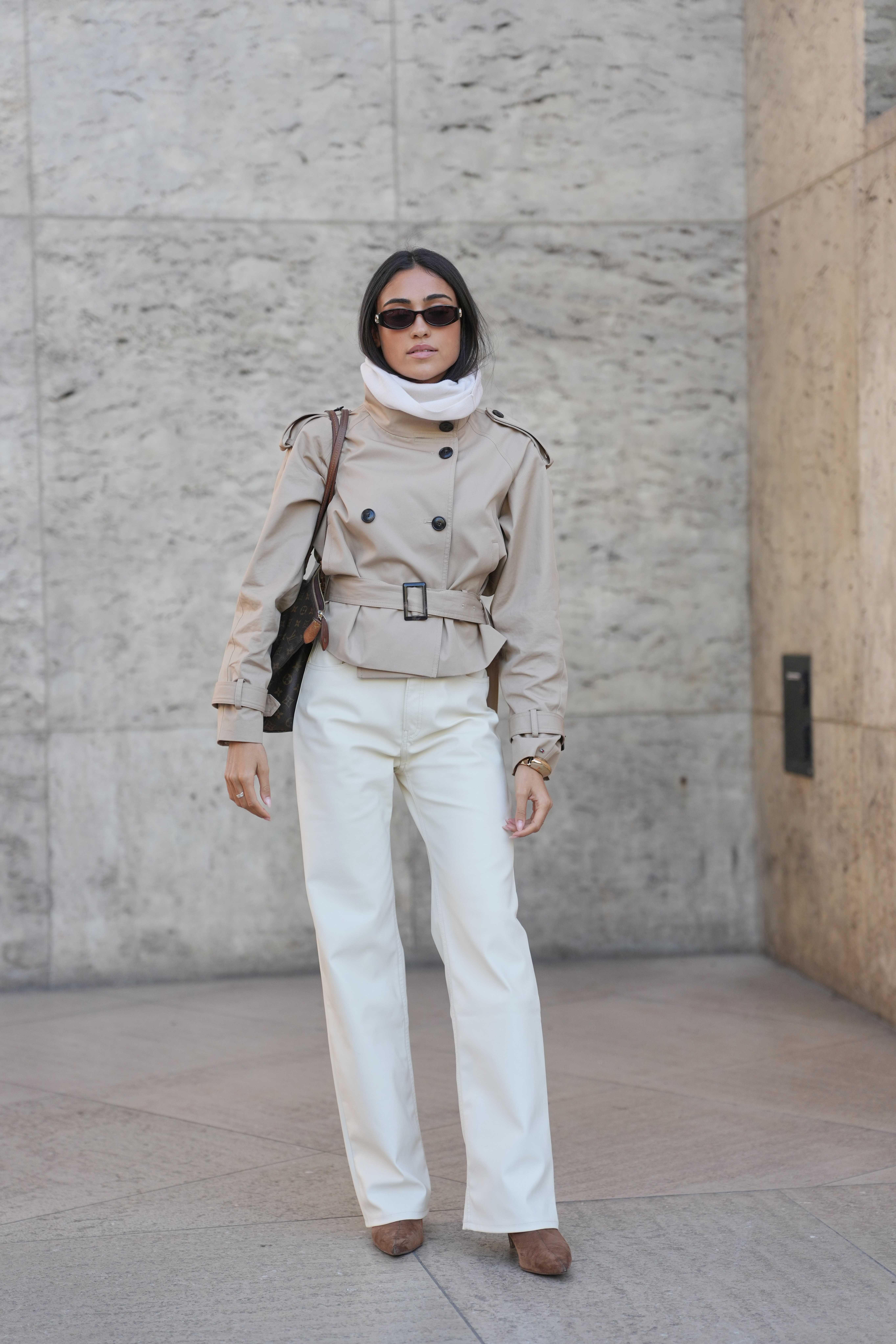 PARIS, FRANCE - OCTOBER 07: A guest wears long straight dark brown hair with a center part, small black oval sunglasses, multiple silver rings and a gold bangle bracelet, a brown monogram coated canvas Louis Vuitton shoulder bag with leather trim, a beige cropped belted trench coat with black buttons and buckle details worn over a white high-neck top, off-white straight-leg trousers, brown suede pointed-toe pumps shoes, outside Ujoh, during Paris Fashion Week - Womenswear Spring Summer 2026, on October 07, 2025 in Paris, France (Photo by Edward Berthelot/Getty Images)
