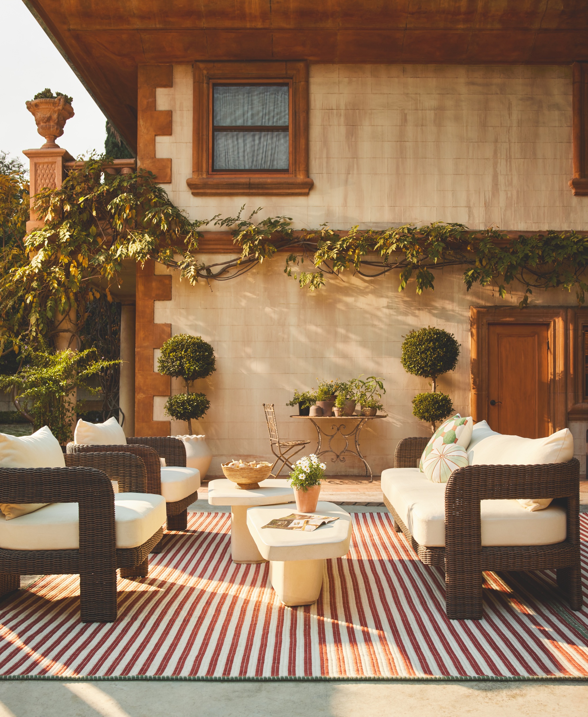 Outdoor living space with a white sofa facing two matching armchairs on a red striped rug and two cream stone coffee tables.