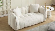 Small cream channel tufted 'boneless' couch styled inside of wood-floor living room area featuring a grey area rug and sculptural side table holding assorted brass objects