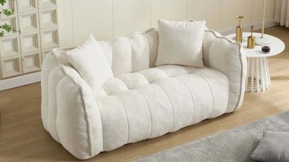 Small cream channel tufted 'boneless' couch styled inside of wood-floor living room area featuring a grey area rug and sculptural side table holding assorted brass objects