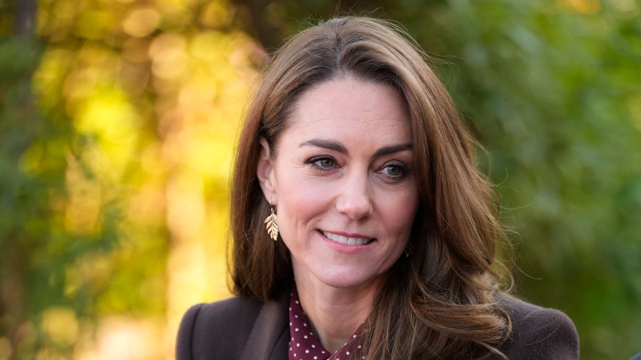 Kate Middleton wearing fern earrings, a brown coat and burgundy dress as she smiles and looks to the side