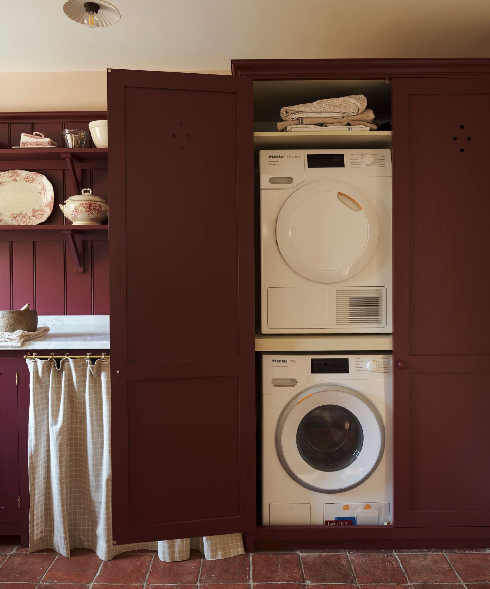 A burgundy laundry room with stacked washer and dryer, next to shelves with patterned ornaments.