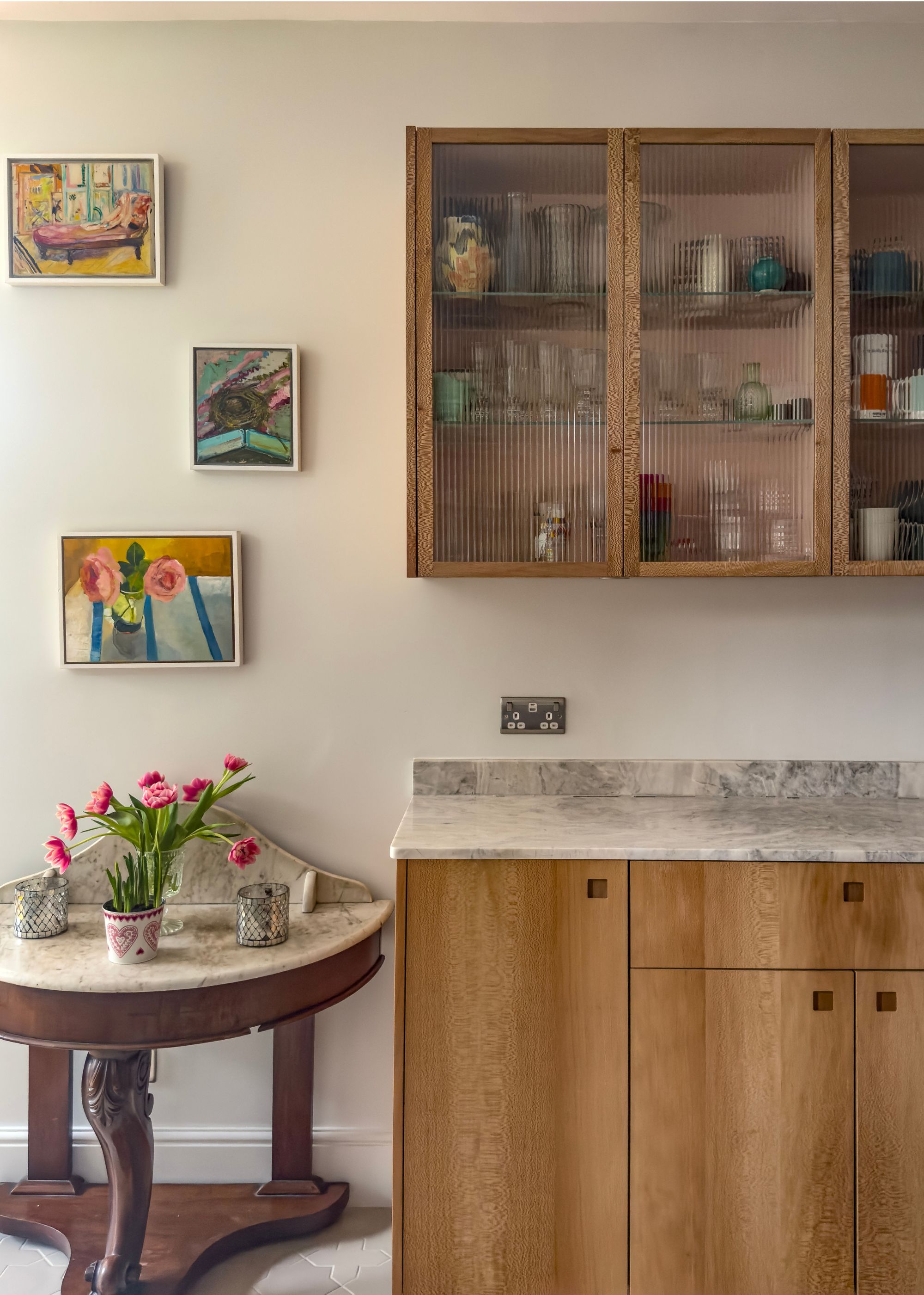 A kitchen with glass fluted cabinets and a tble on the side with flowers and a plant pot. Above the table are three framed art pieces.