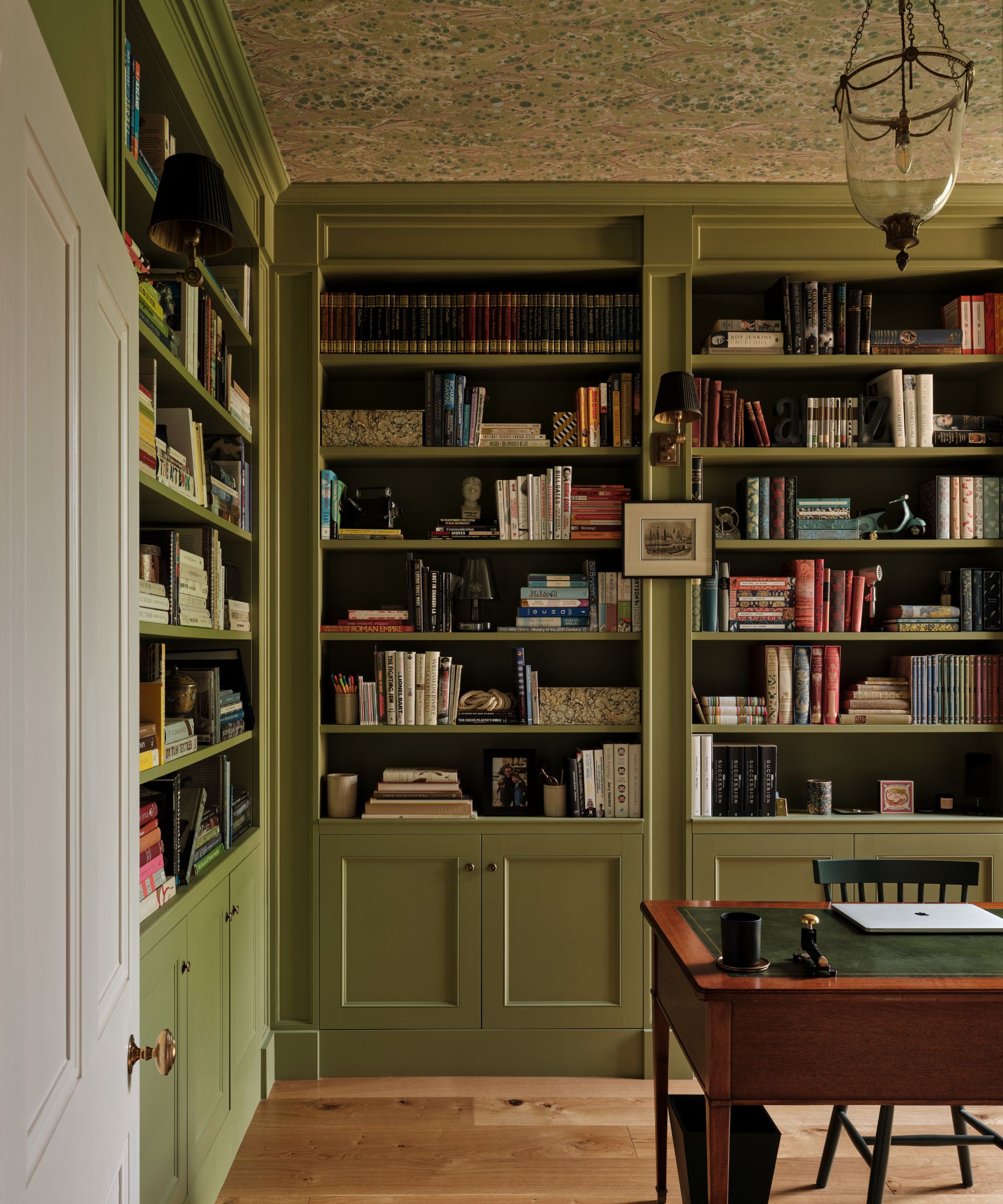 Study with green-painted built-in bookcases covered in books, a vintage wooden writing desk, a wallpapered ceiling, and an antique lantern pendant light.