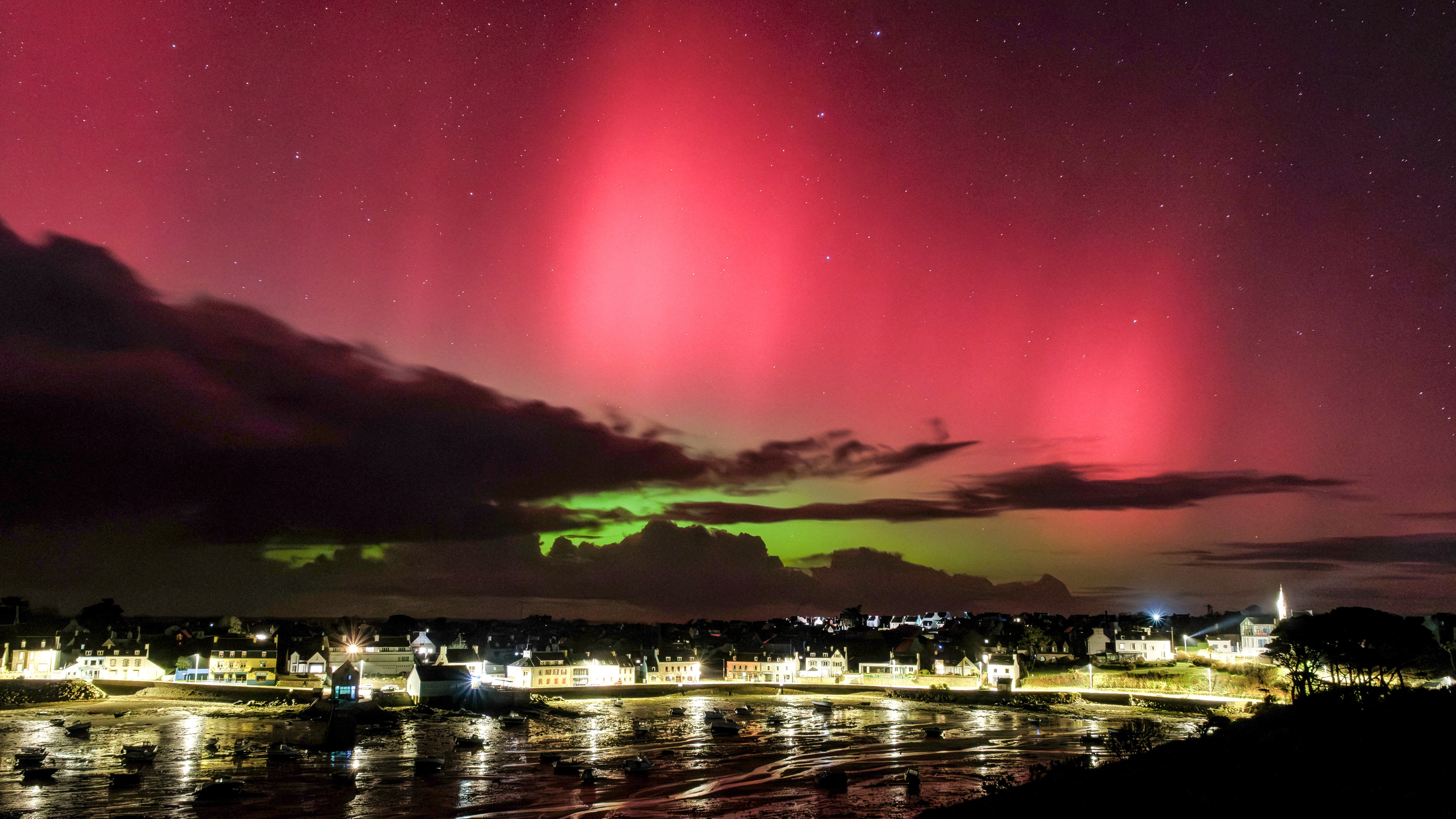 vibrant red aurora fills the sky with some bright green aurora below. there are lights from houses that line the bay reflected in the water below.
