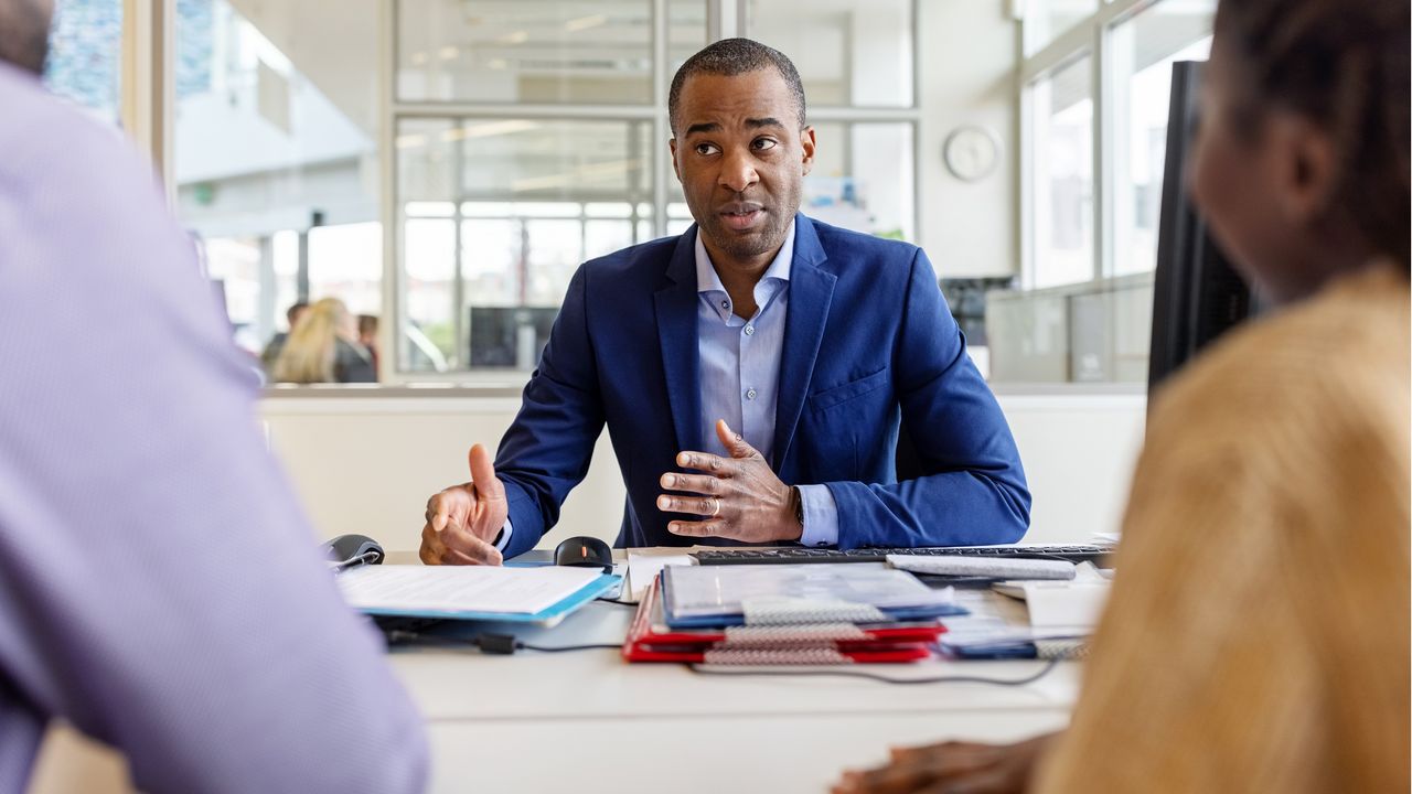 A man sits behind a desk and talks to two customers.