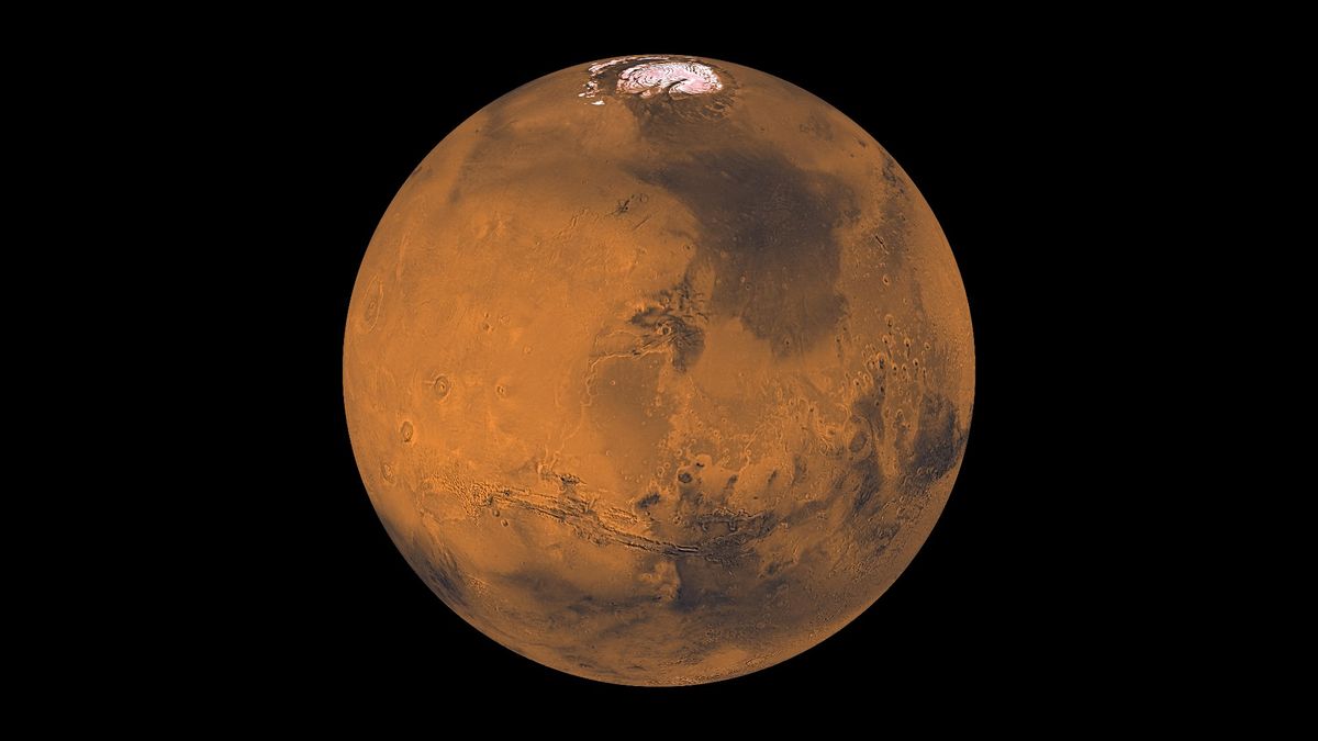 Global color view of Mars. A large, round, rusty-red planet. There are numerous craters all over on the planet. There is a small white circle at the top (ice cap). In the top-right quadrant of the planet there is a very large darker patch. On the bottom half there is what appears to be a long canyon that is in the shape of a smile.