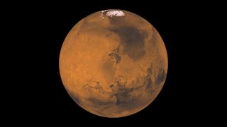 Global color view of Mars. A large, round, rusty-red planet. There are numerous craters all over on the planet. There is a small white circle at the top (ice cap). In the top-right quadrant of the planet there is a very large darker patch. On the bottom half there is what appears to be a long canyon that is in the shape of a smile.