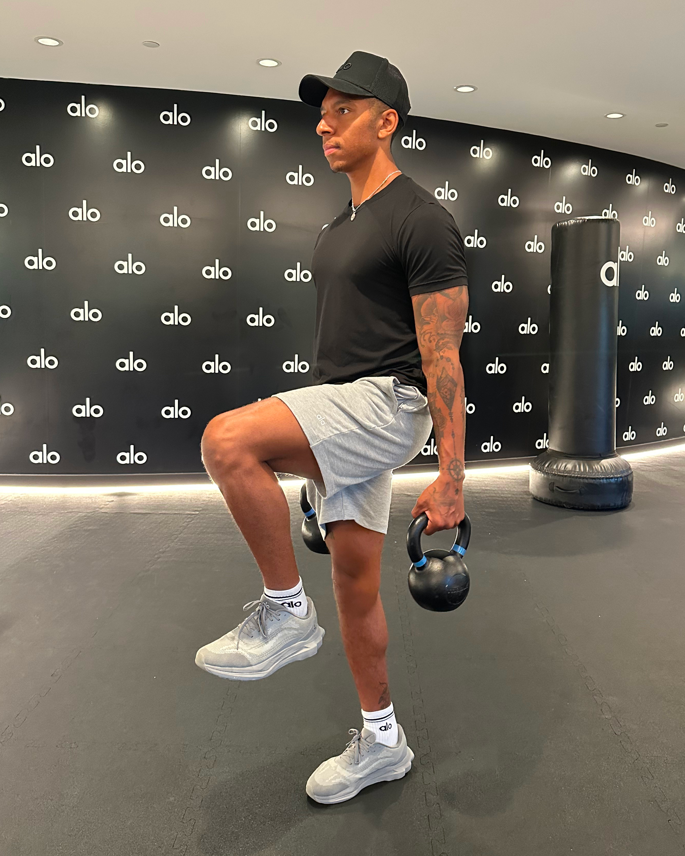 Man in black T-shirt, black cap, white shorts and white trainers demonstrates exercise
