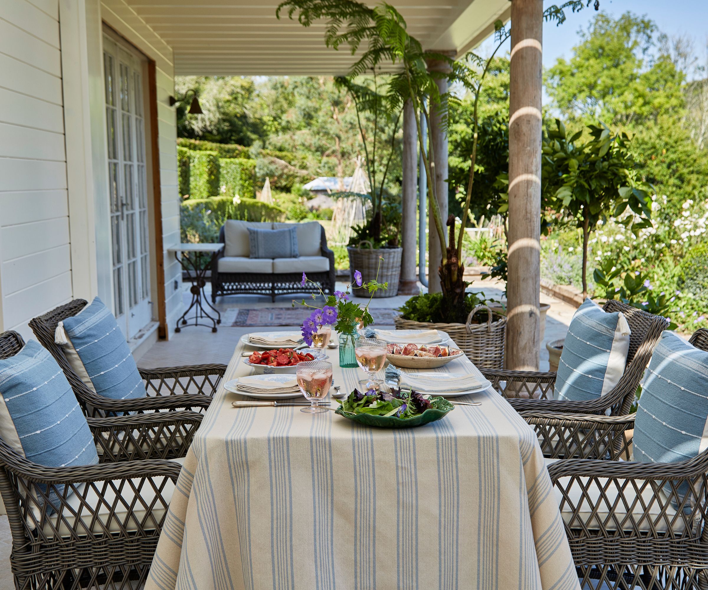 Nancy Meyers style outdoor porch with a wicker dining set laid with a coastal tablescape with a striped tablecloth and sharing dishes