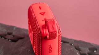 The corner loop of a red JBL Go 4 pictured against a pink background. It is sitting on a dark stone surface with puddles of water around it.