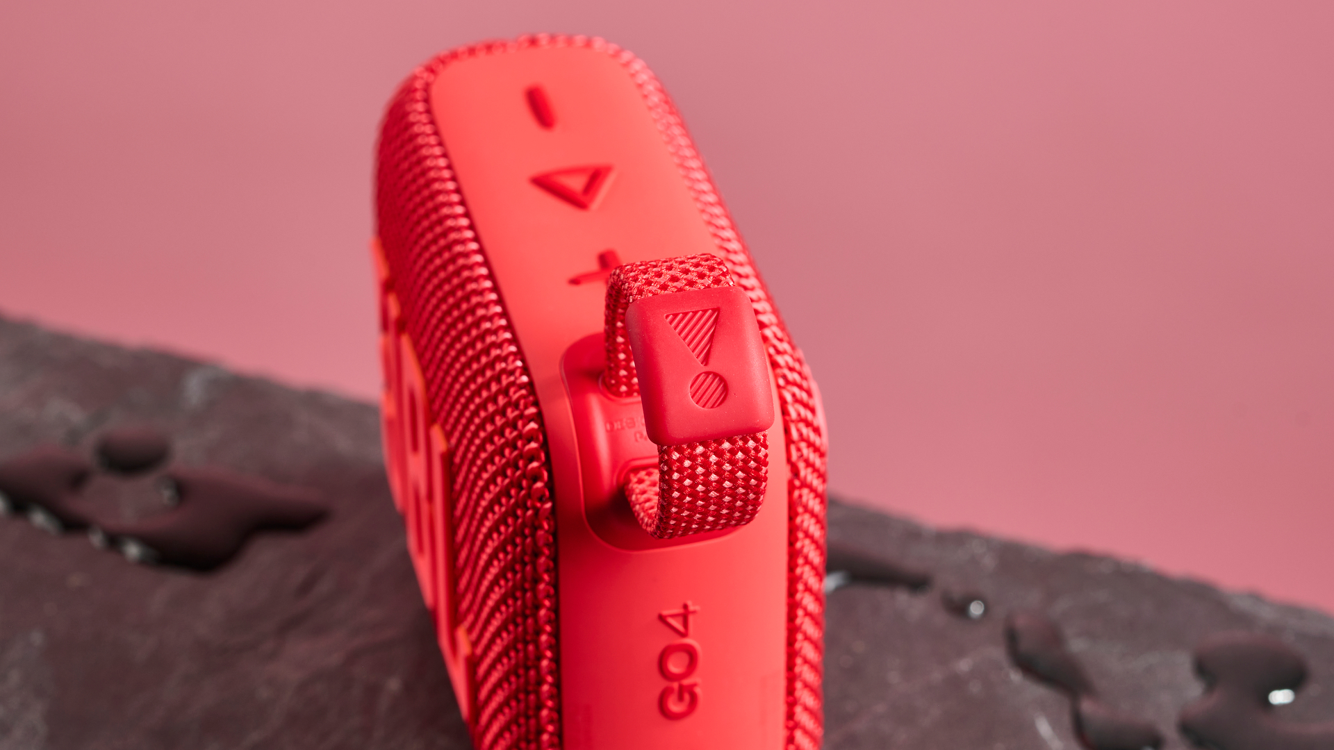 The corner loop of a red JBL Go 4 pictured against a pink background. It is sitting on a dark stone surface with puddles of water around it.