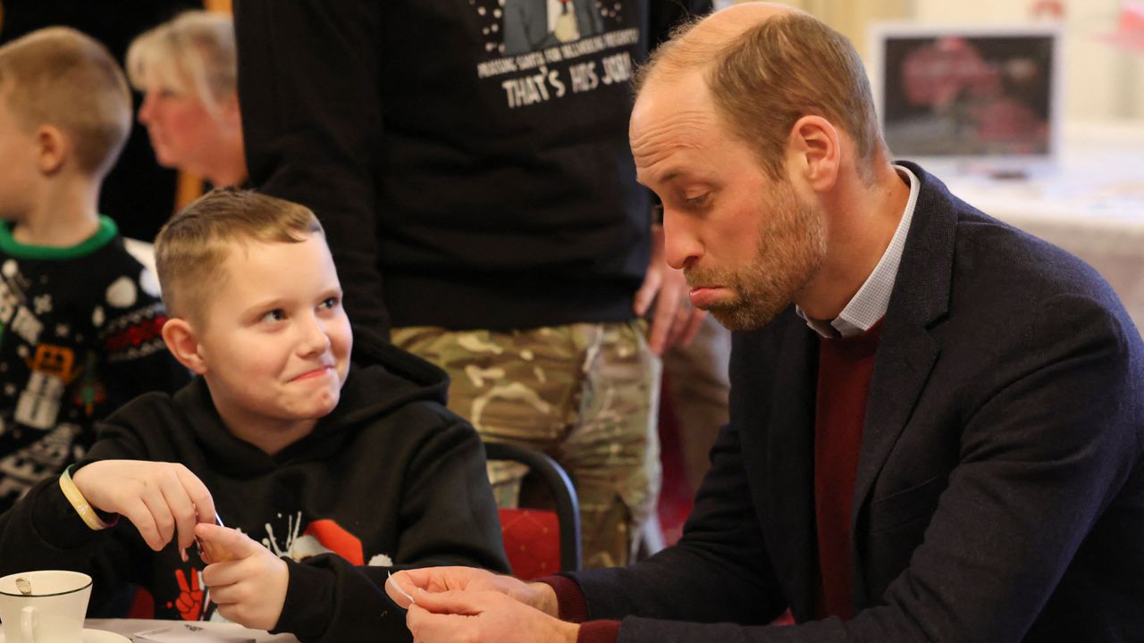 Prince William sitting at a table with a little boy and making a funny pouting expression looking at a paper