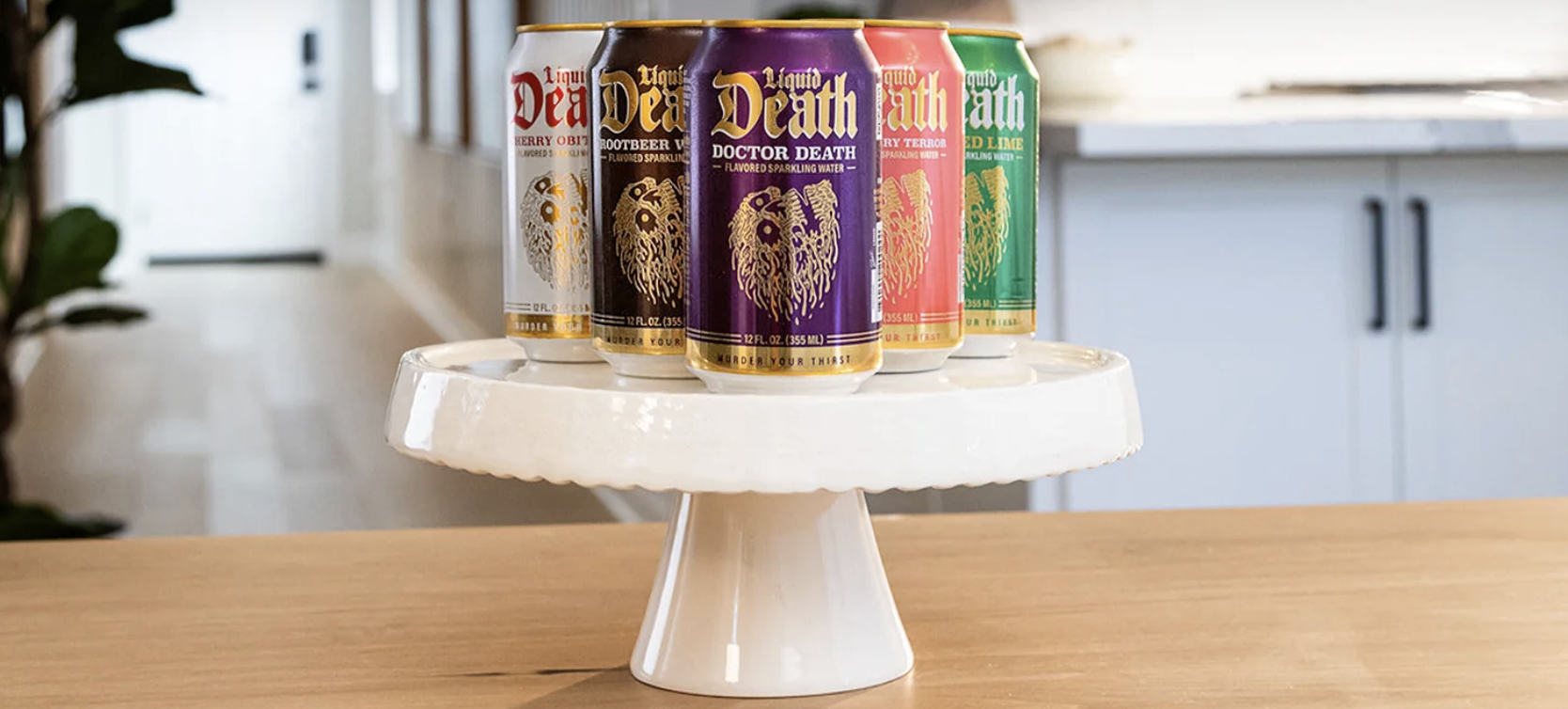 Five colorful cans of Liquid Death flavored sparkling water are displayed in a row on a white ceramic cake stand atop a wooden counter.