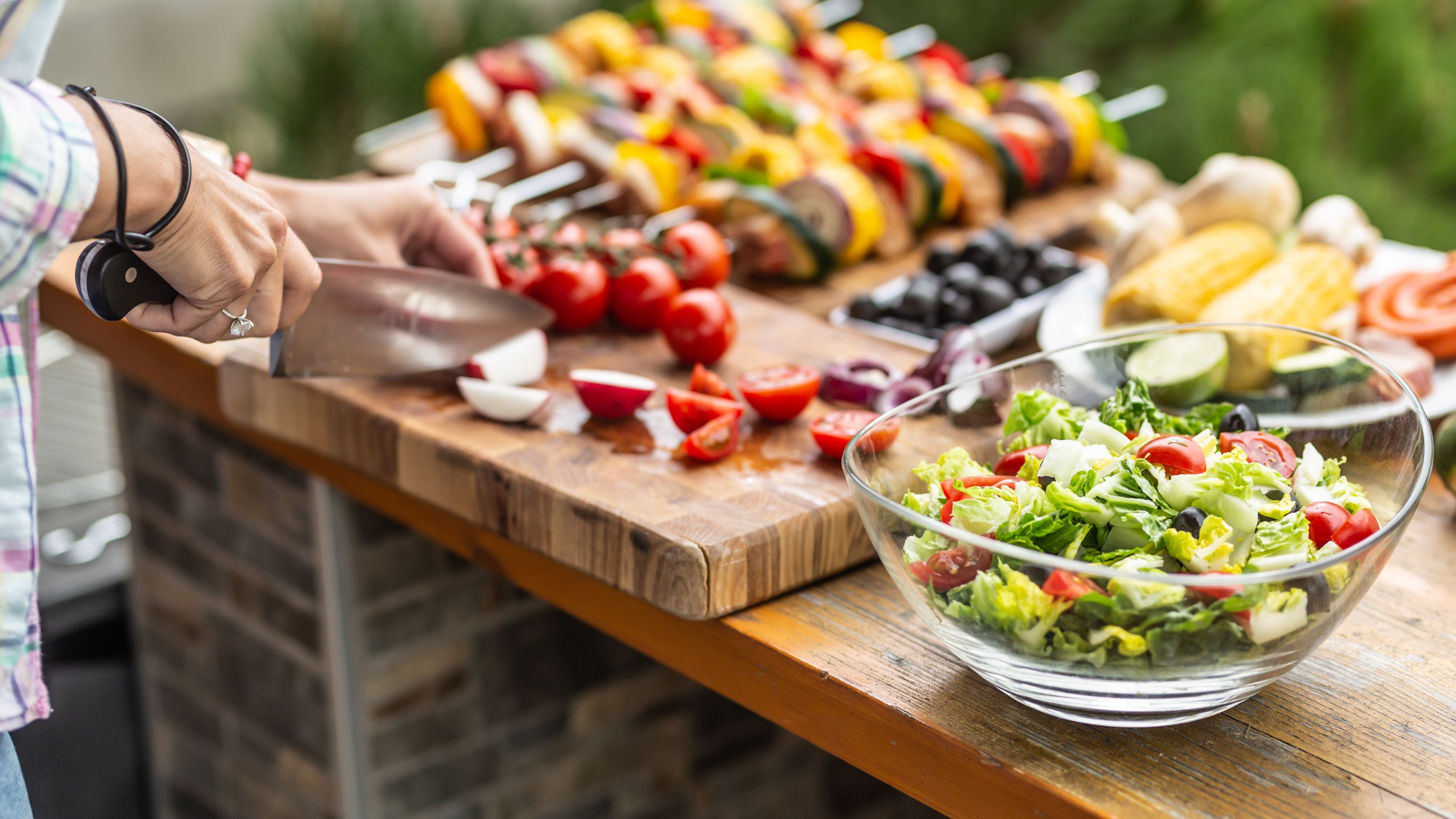 Garden table full of food ready for a barbeque party with a salad in the front and a woman cutting vegetable aside.