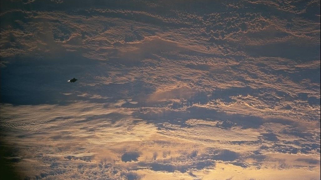 images captured from space show earth covered in clouds below and a small black object visible towards the left of the image.