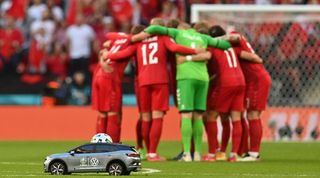 Denmark players in a huddle ahead of their match against England at Euro 2020 as the Volkswagen remote control car brings the match ball onto the pitch in the foreground.