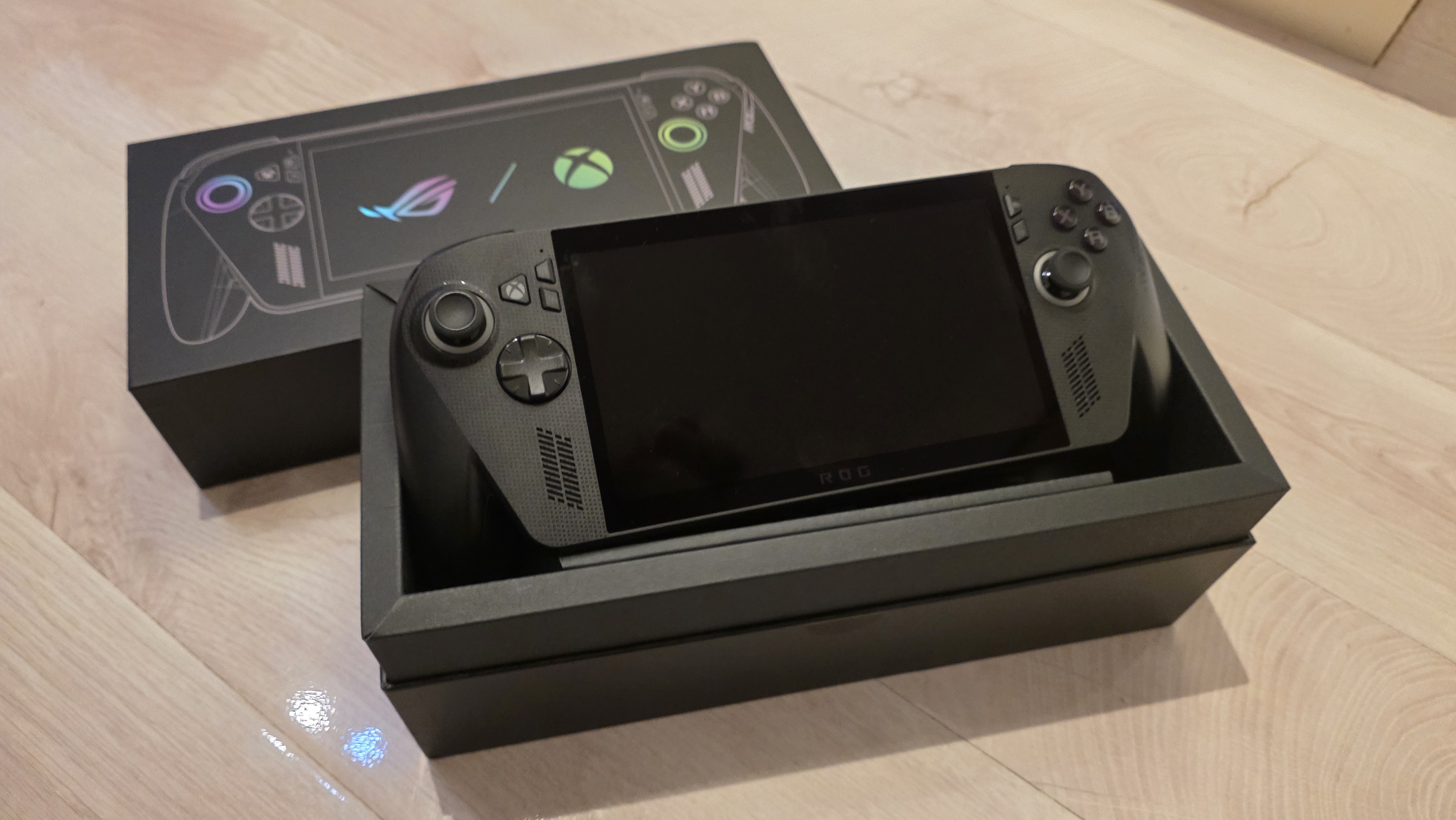 A black ROG gaming handheld console sits in an open black box on a wooden surface. The box displays controller graphics with vibrant icons.