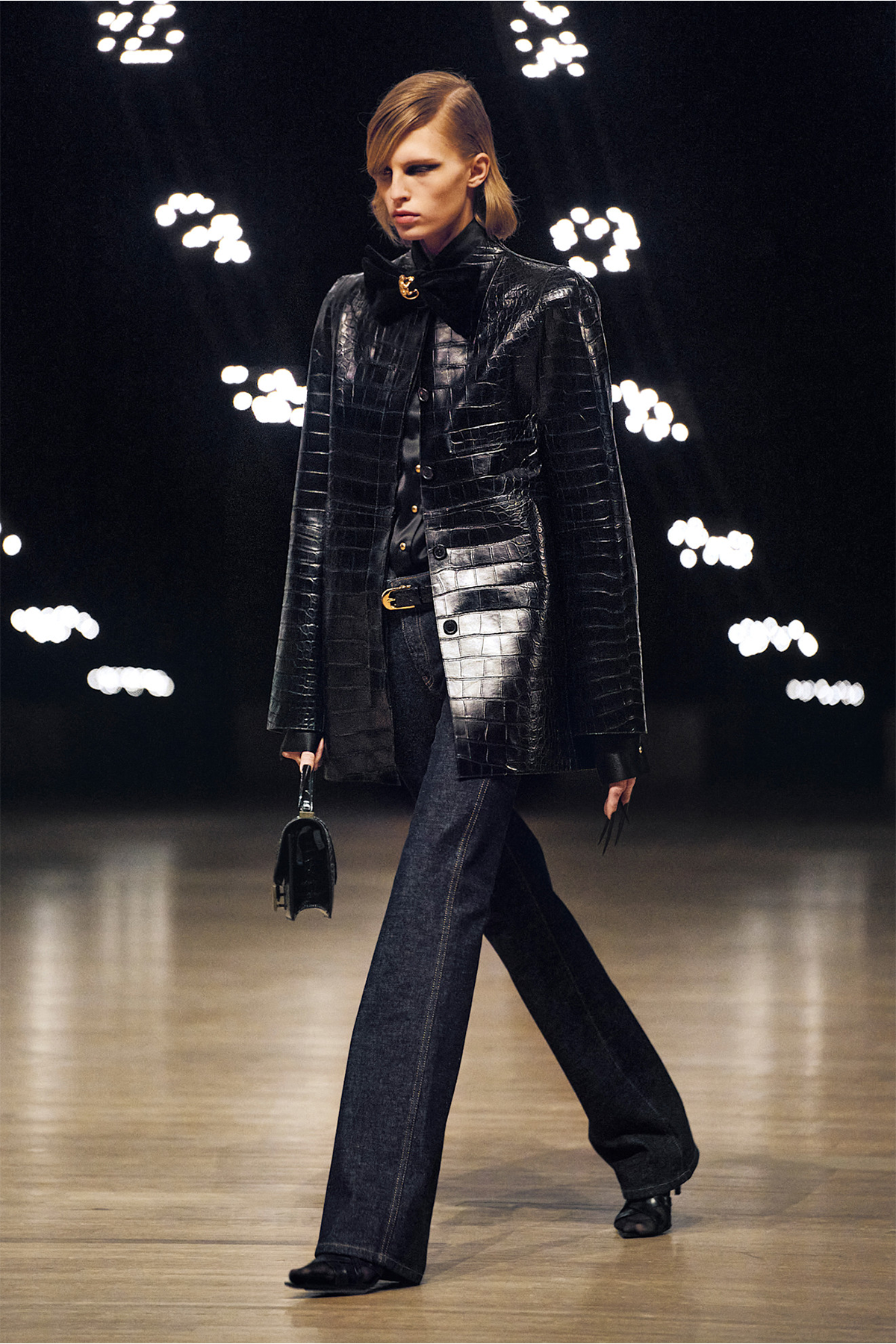 A model on the Khaite F/W 2026 runway wearing indigo straight jeans, a croc leather coat, black boots and top-handle bag