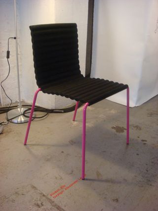 Concrete floor, white walls, chair with black rib design seat and four pink legs, silver lamp stand, brown piping up wall and along floor