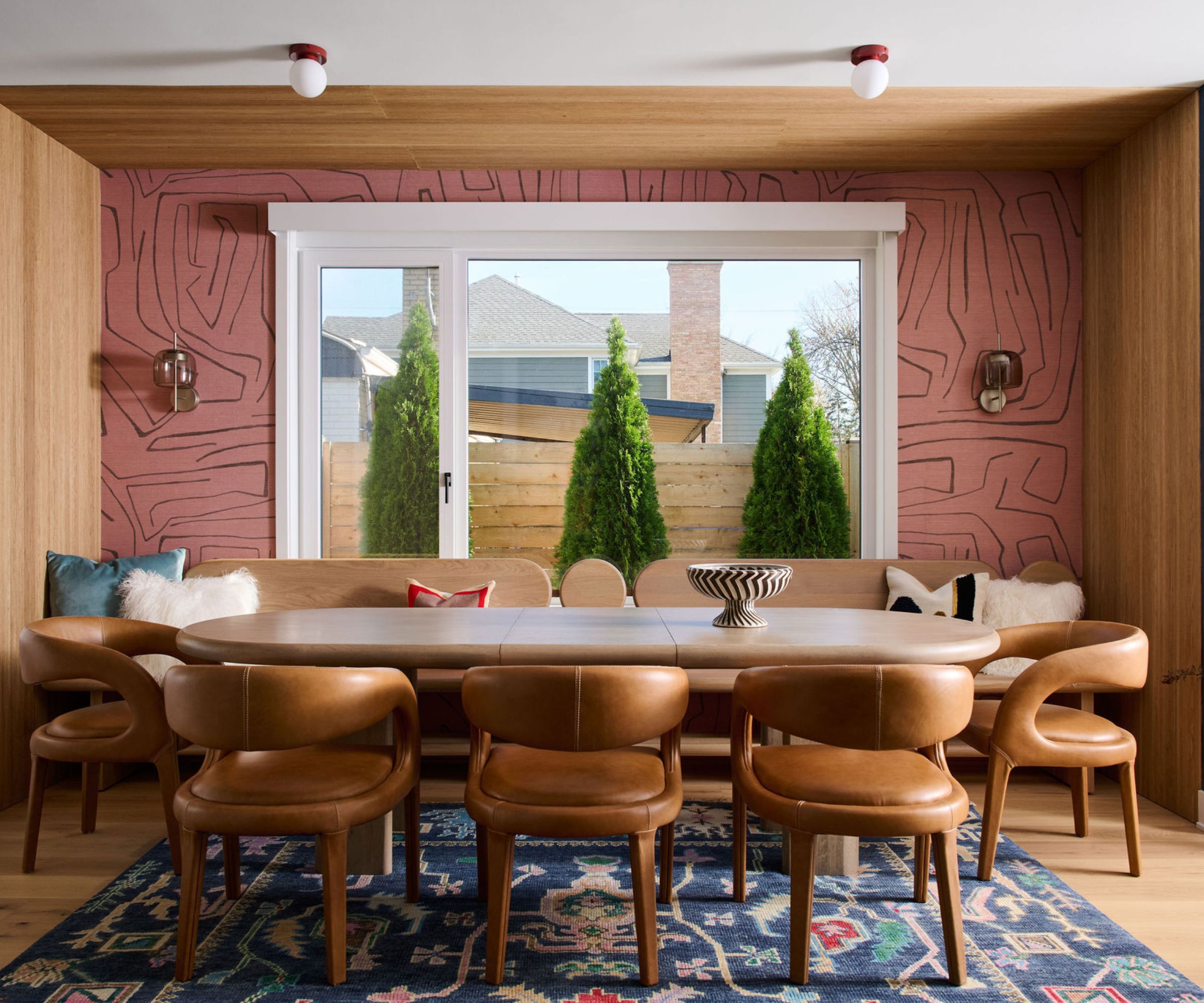 Dining room with pink abstract wallpaper, wooden long curved dining table, brown leather chairs, banquette, and oushak rug