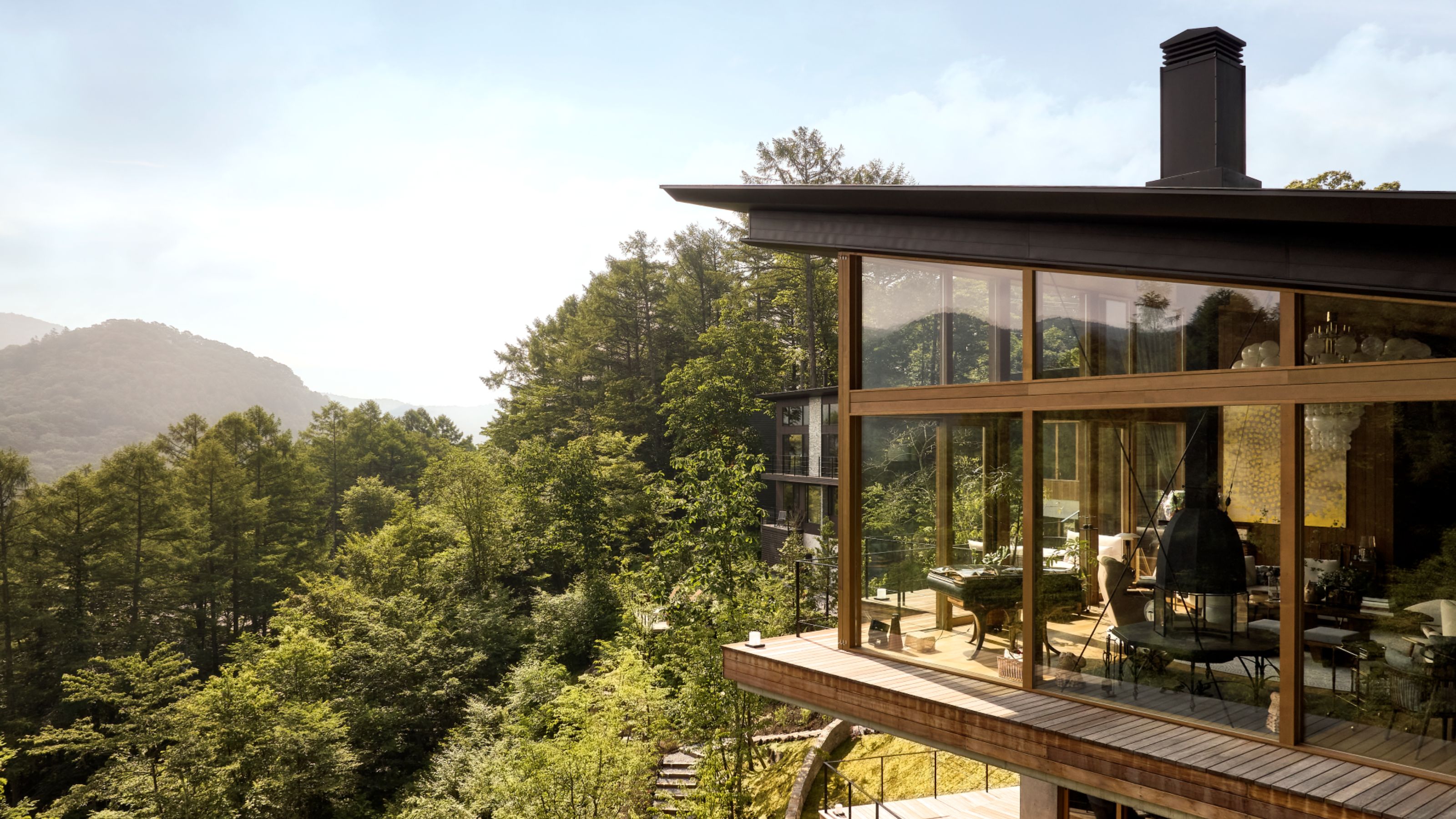 A grand Japanese home angled towards the surrounding forest and mountains with large wood-paneled glass windows all around and a sloped black roof with a black chimney breast