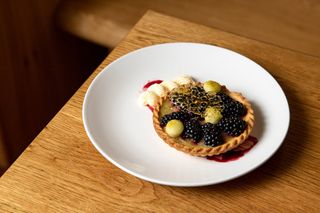 A wooden table holds a white plate filled with a creatively presented fruit tart dessert, covered in honeycomb.