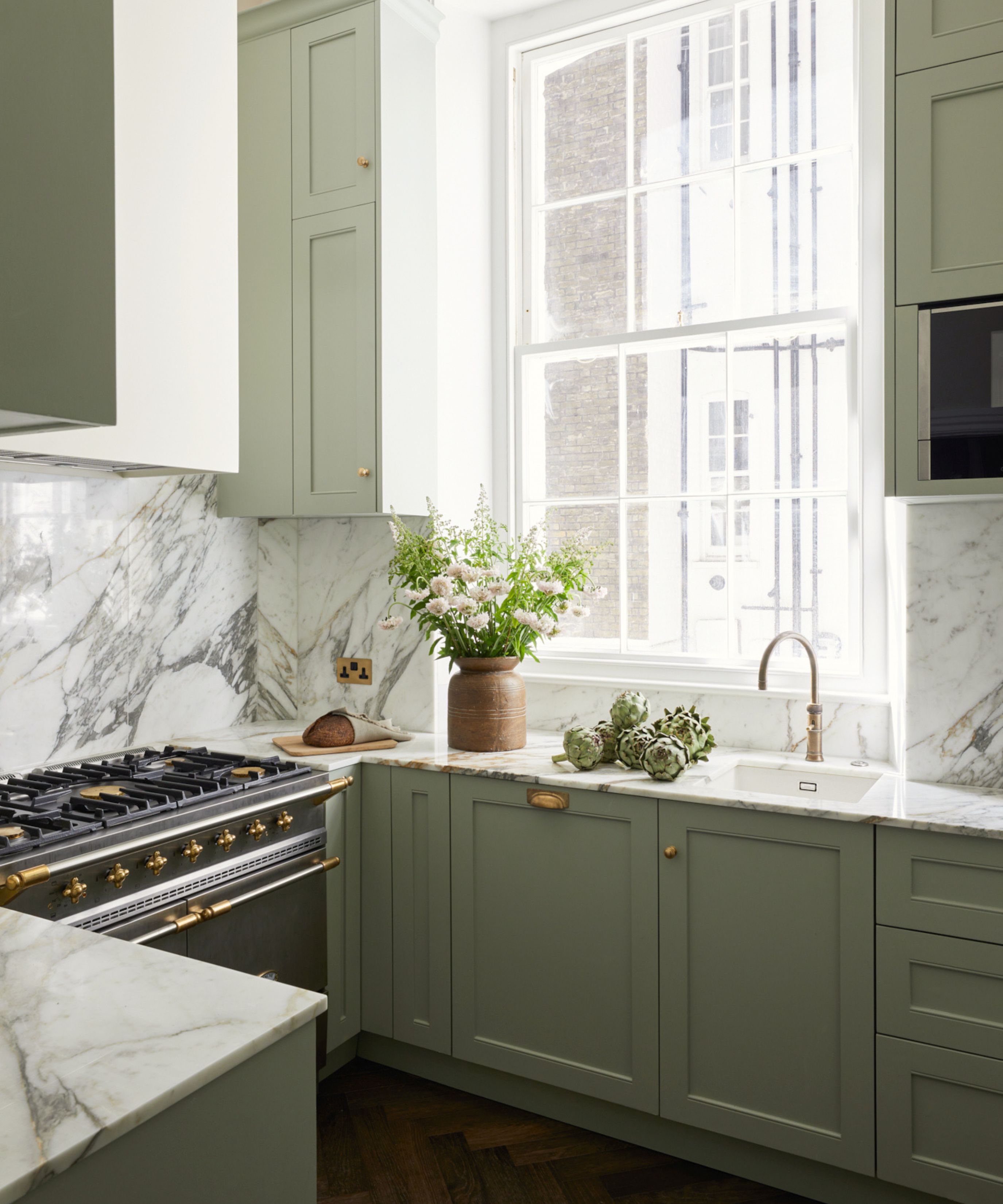 A bright, airy kitchen with sage green shaker-style cabinets and heavy-veined white marble backsplashes. Sunlight streams through a large sash window onto a countertop holding a vase of white flowers and fresh artichokes.