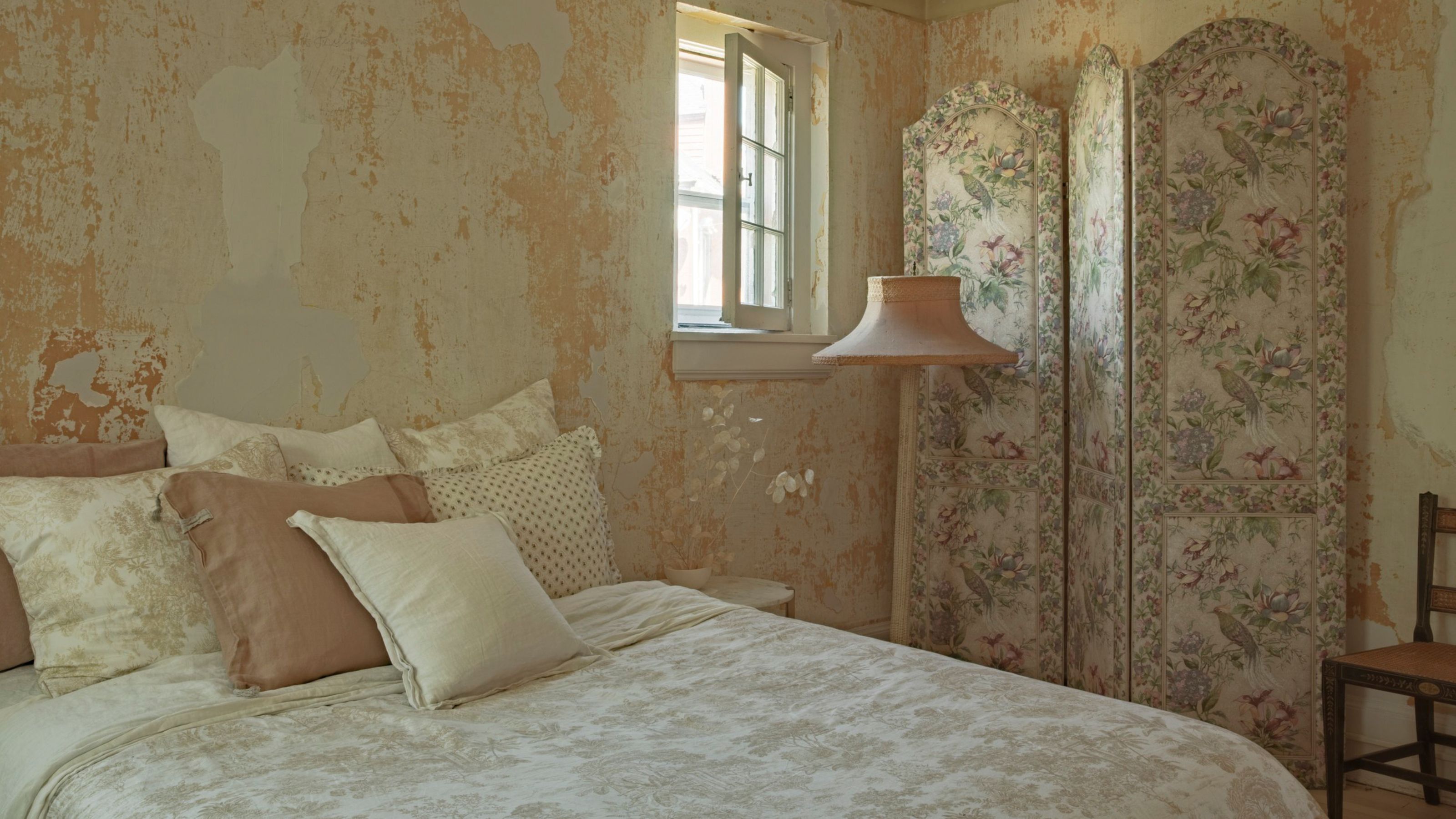 a yellow plaster peeling historic bedroom with vintage floral screen and layered neutral linens