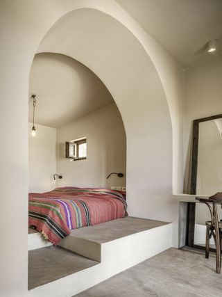 A bedroom with cream walls and grey flooring featuring an arch with platforms that leads to the double bed covered with pink covers with striped. About the bed is hanging lighting and small open window